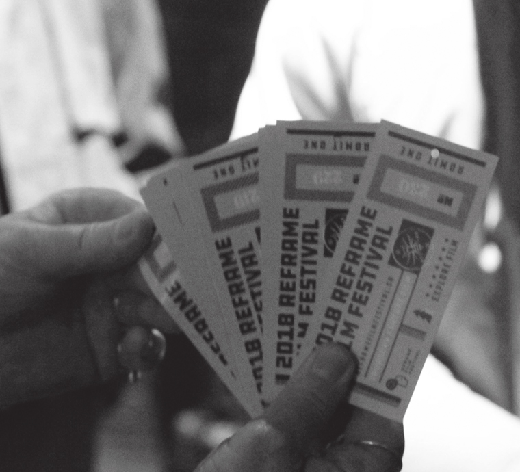 A grayscale image of a person's hand holding several tickets. The tickets are in focus and display text indicating they are for a festival called 'Reframe,' with other text details blurred and unreadable. The background is out of focus, with the silhouette of a person discernible.